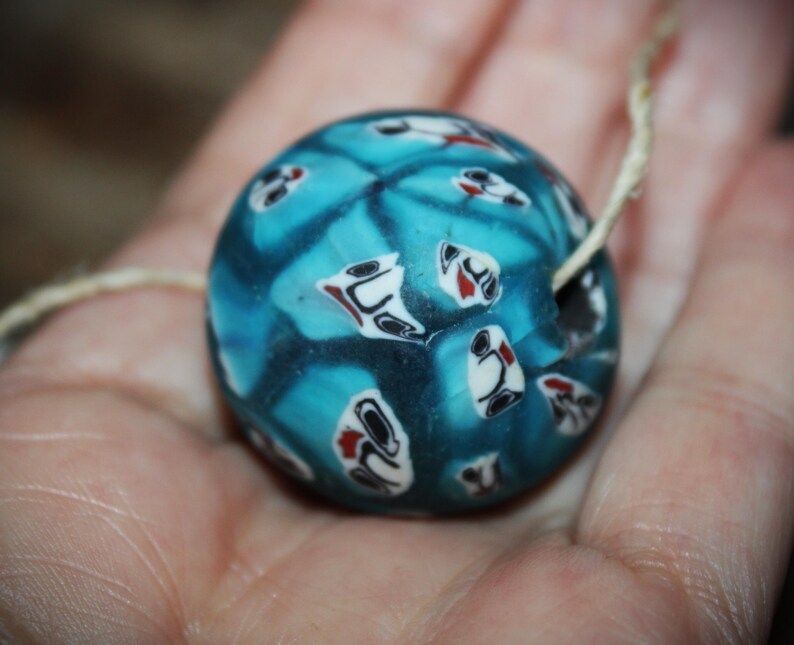 K&ouml;nnte beinhalten: Eine runde, blaue Glasperle mit einem dekorativen Muster aus wei&szlig;en, schwarzen und roten Formen. Die Perle hat ein Loch zum Auff&auml;deln und wird in einer Hand gehalten. Die Perle hat einen Durchmesser von etwa 2,5 cm.