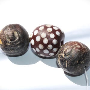Puede incluir: Tres cuentas de color marrón oscuro con texturas visibles. Una cuenta es de color granate oscuro con lunares blancos. Las cuentas están ensartadas en un hilo transparente, lo que sugiere que son para hacer joyas o manualidades.