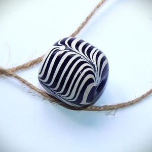 May include: A dark blue and white striped ceramic bead with a swirl pattern. The bead is strung on a natural twine cord.