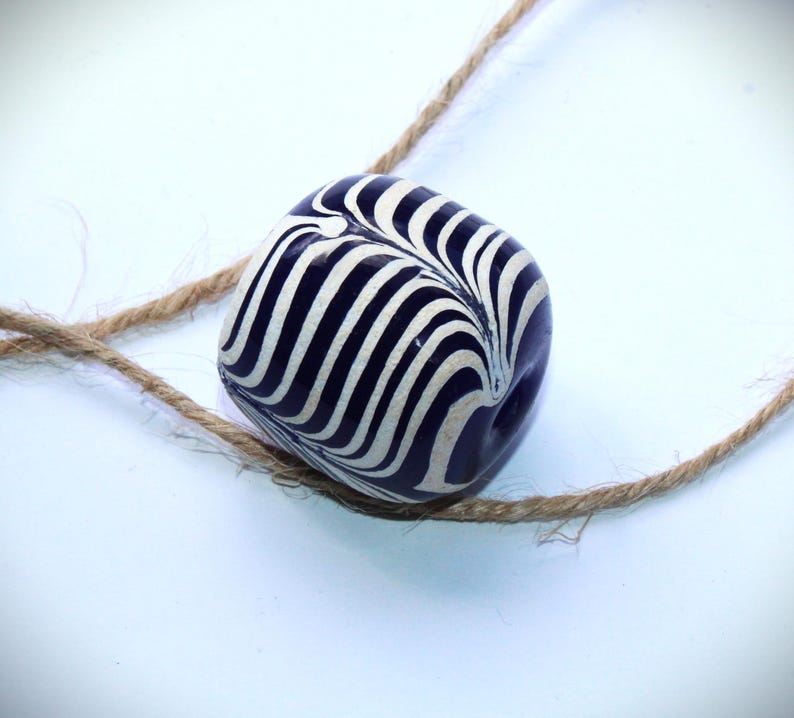 May include: A dark blue and white striped ceramic bead with a swirl pattern. The bead is strung on a natural twine cord.