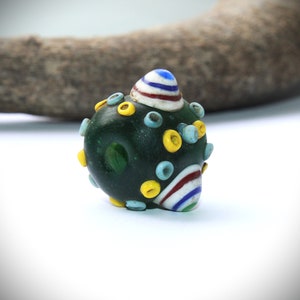 May include: A dark green glass bead with colorful accents. The bead features yellow, light blue, and white circular designs. A striped pattern in red, white, and blue adorns one end, and a striped top in blue, white, and red is on the other end.