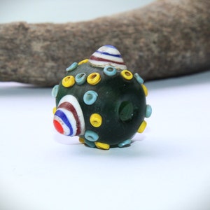 May include: A dark green glass bead with colorful accents. The bead features a rounded shape with yellow, light blue, and white circular designs. A striped cone shape is on top, with red, white, and blue stripes. The bead has a hole through the center.