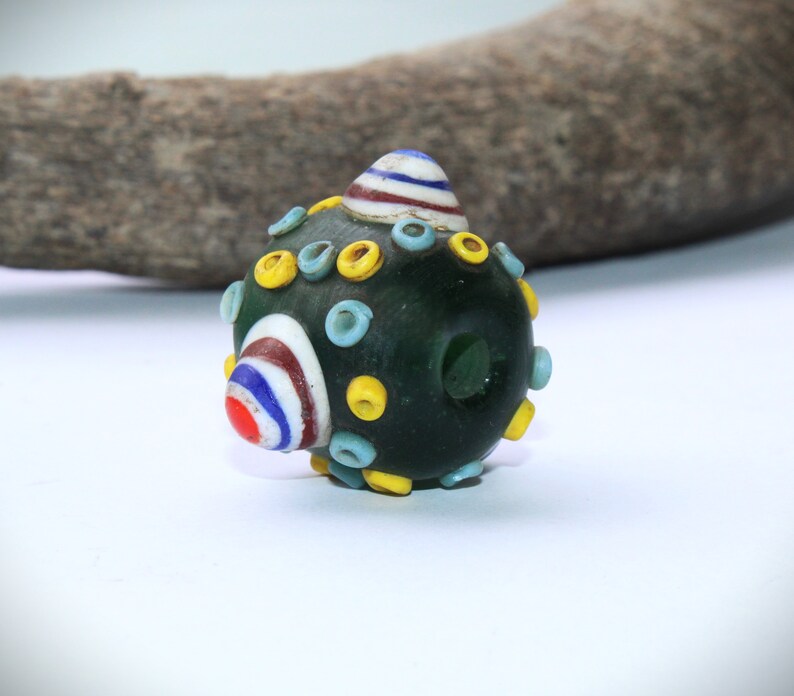 May include: A dark green glass bead with colorful accents. The bead features a rounded shape with yellow, light blue, and white circular designs. A striped cone shape is on top, with red, white, and blue stripes. The bead has a hole through the center.