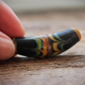 Puede incluir: Primer plano de una cuenta de vidrio colorida con remolinos negros, amarillos, naranjas y verdes. La cuenta está siendo sostenida por un dedo.