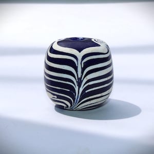 Könnte beinhalten: Eine blaue und weiße Glasperle mit einem wirbelnden Muster. Die Perle ist rund und hat ein Loch in der Mitte.