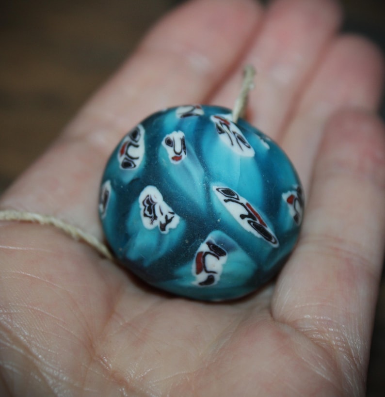 K&ouml;nnte beinhalten: Eine runde, blaue Glasperle mit wei&szlig;en, schwarzen und roten dekorativen Mustern. Die Perle hat eine Schnur und wird in einer Hand gehalten. Das Design der Perle ist eine Mischung aus abstrakten Formen und floralen Elementen.