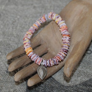Puede incluir: Un brazalete colorido hecho de pequeñas cuentas planas en forma de concha en tonos naranja, morado y amarillo. Un dije de hoja plateada cuelga del brazalete. El brazalete se muestra en una mano de madera.
