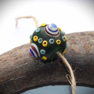 May include: A dark green glass bead with colorful accents. The bead features a design of white, red, and blue stripes, along with yellow and light blue circles. A natural twine string is attached, suggesting it could be used for jewelry or crafts.