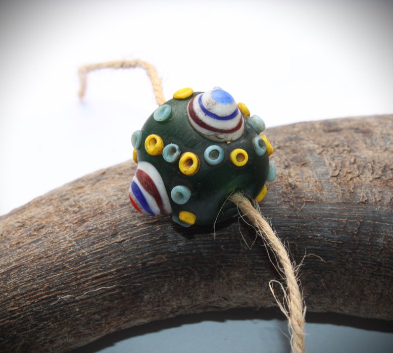 May include: A dark green glass bead with colorful accents. The bead features a design of white, red, and blue stripes, along with yellow and light blue circles. A natural twine string is attached, suggesting it could be used for jewelry or crafts.