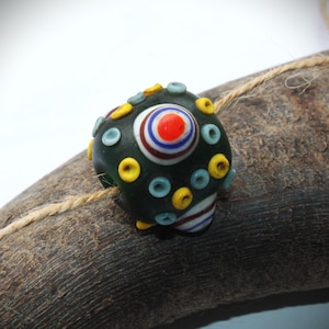 Könnte beinhalten: Eine dunkelgrüne Glasperle mit einem zentralen Design aus konzentrischen Kreisen in Rot, Weiß und Blau sowie einem gelb-blauen Muster. Die Perle ist mit gelben und blauen Akzenten verziert und liegt auf einem Holzstück mit natürlichem Bindfaden.