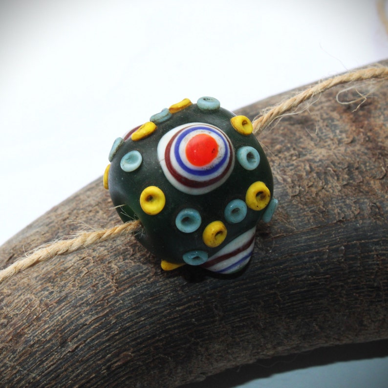 May include: A dark green glass bead with a central design of concentric circles in red, white, blue, and a yellow and blue pattern. The bead is adorned with yellow and blue accents and rests on a piece of wood with a natural twine.