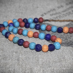 Könnte beinhalten: Eine Halskette mit einer Kette aus farbenfrohen, strukturierten Perlen in Blau-, Rot- und Orangetönen. Die Perlen sind auf einer naturfarbenen Kordel aufgefädelt. Die Halskette wird auf einer strukturierten, grauen Oberfläche präsentiert.