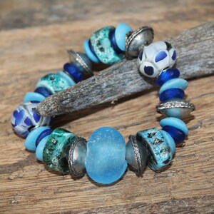 Peut inclure: Un bracelet en perles bleues et argent avec une grosse perle en verre bleu clair au centre. Le bracelet est composé de différentes perles, notamment turquoise, bleues et argentées.