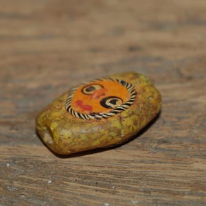 Puede incluir: Una cuenta rectangular de color amarillo texturizado con una incrustaci&oacute;n circular naranja central que presenta un rostro pintado a mano. El rostro tiene ojos delineados en negro, una peque&ntilde;a nariz naranja y labios rojos, rodeado por un borde a rayas blancas y negras.
