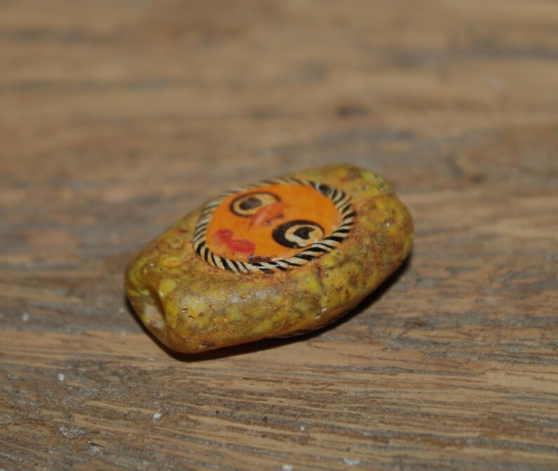 Puede incluir: Una cuenta rectangular de color amarillo texturizado con una incrustaci&oacute;n circular naranja central que presenta un rostro pintado a mano. El rostro tiene ojos delineados en negro, una peque&ntilde;a nariz naranja y labios rojos, rodeado por un borde a rayas blancas y negras.