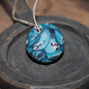 K&ouml;nnte beinhalten: Eine runde, blaue Perle mit einem wirbelnden Muster aus wei&szlig;en, roten und schwarzen Formen. Die Perle ist an einem d&uuml;nnen, naturfarbenen Faden aufgeh&auml;ngt. Die Perle liegt auf einer dunkelgrauen Holzoberfl&auml;che.