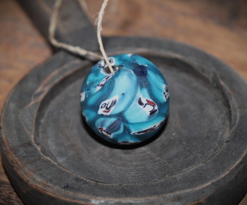 K&ouml;nnte beinhalten: Eine runde, blaue Perle mit einem wirbelnden Muster aus wei&szlig;en, roten und schwarzen Formen. Die Perle ist an einem d&uuml;nnen, naturfarbenen Faden aufgeh&auml;ngt. Die Perle liegt auf einer dunkelgrauen Holzoberfl&auml;che.