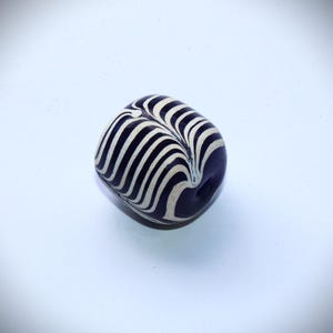 May include: A dark blue and white striped glass bead. The bead has a swirl pattern and is oval shaped.