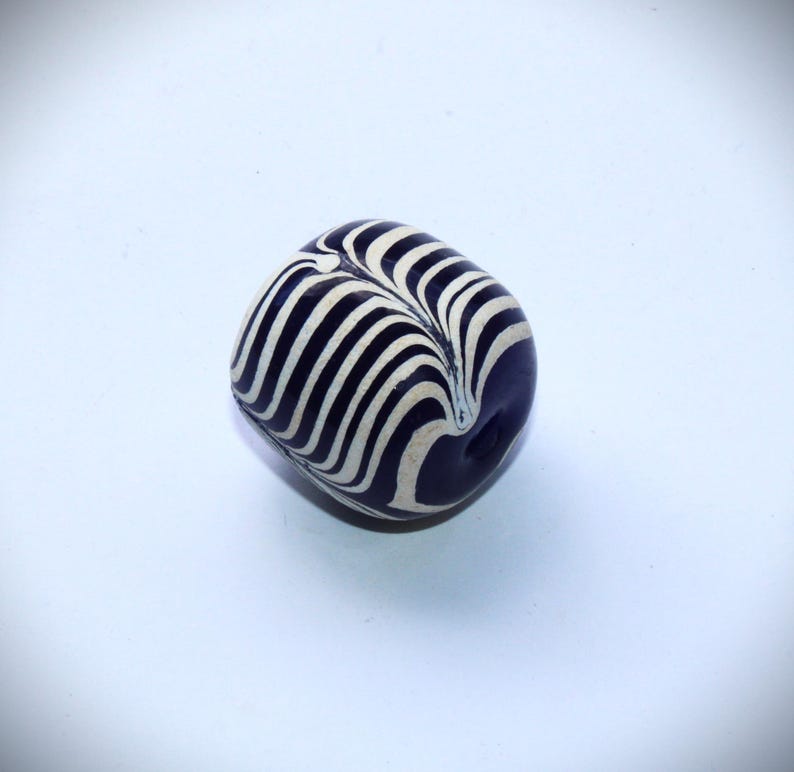 May include: A dark blue and white striped glass bead. The bead has a swirl pattern and is oval shaped.
