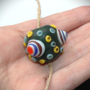 May include: A dark green glass bead with colorful circular designs in red, white, blue, and yellow. The bead is strung on a natural fiber cord. The bead has a unique, handcrafted appearance.
