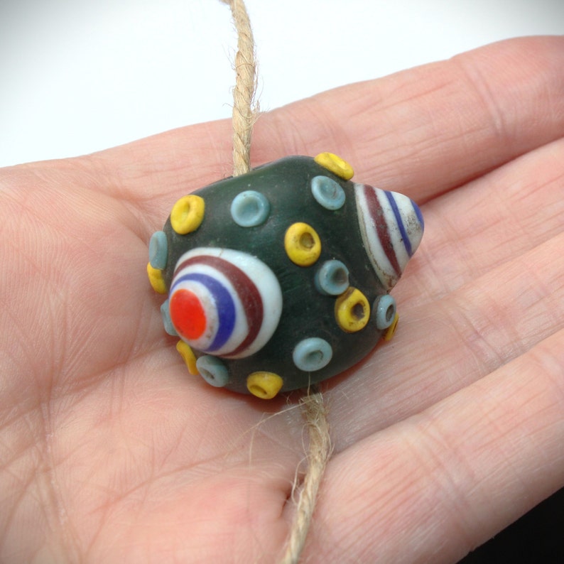 May include: A dark green glass bead with colorful circular designs in red, white, blue, and yellow. The bead is strung on a natural fiber cord. The bead has a unique, handcrafted appearance.
