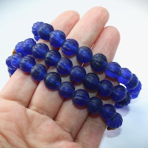 Könnte beinhalten: Eine Strähne blauer Glasperlen mit einer strukturierten, kürbisartigen Form. Die Perlen sind auf einer Schnur aufgefädelt und werden auf einer Hand gezeigt.