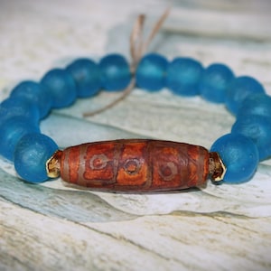 May include: A handmade bracelet featuring large, round, light blue beads. The focal bead is a brown, patterned barrel bead with gold-colored end caps. The bracelet is strung on a natural-colored cord.