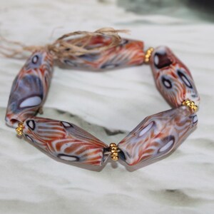 Puede incluir: Una pulsera con cuentas oblongas con un diseño veteado en tonos naranja, blanco y gris. Las cuentas están unidas con espaciadores dorados y un cierre de cuerda natural. Un accesorio único.
