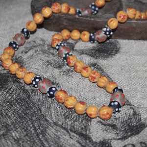 Könnte beinhalten: Eine Halskette mit einer Mischung aus orangefarbenen und gemusterten Perlen. Die orangefarbenen Perlen haben eine strukturierte Oberfläche mit roten Sprenkeln. Die Halskette enthält auch klare Perlen mit roten Punkten und dunkelblaue Perlen mit weißen Punkten.