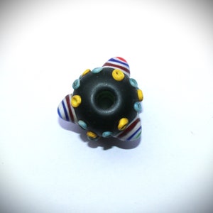 May include: A handmade glass bead with a dark center and colorful accents. The bead has a triangular shape with striped sections in red, white, and blue. Small yellow and blue dots are evenly spaced around the dark center.