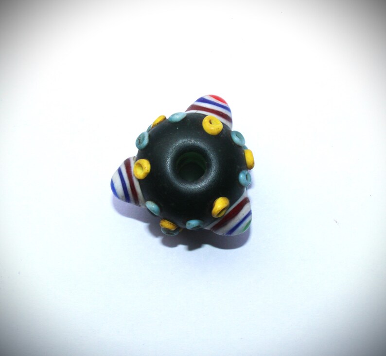 May include: A handmade glass bead with a dark center and colorful accents. The bead has a triangular shape with striped sections in red, white, and blue. Small yellow and blue dots are evenly spaced around the dark center.