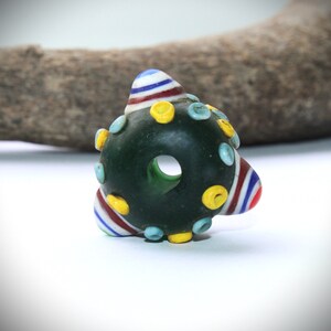 May include: Dark green glass bead with a central hole, adorned with colorful accents. The bead features striped conical shapes in red, white, and blue, along with yellow and light blue circular embellishments. The bead is set against a white background.