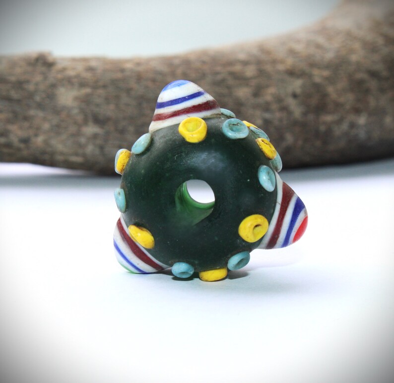 May include: Dark green glass bead with a central hole, adorned with colorful accents. The bead features striped conical shapes in red, white, and blue, along with yellow and light blue circular embellishments. The bead is set against a white background.