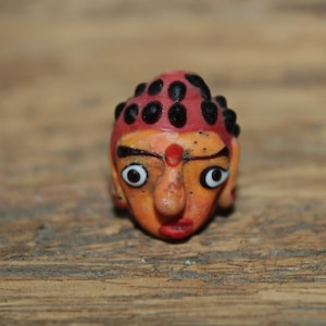 May include: A small, hand-painted figurine of a stylized face. The face is orange with red lips and a red dot on the forehead. The head is topped with a red cap with black dots. The eyes are large and white with black pupils.