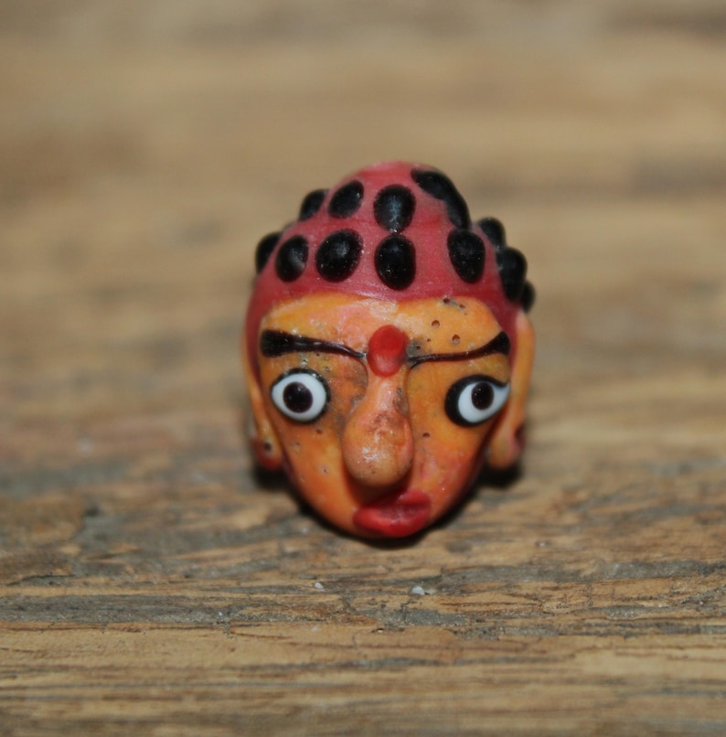 Może przedstawiać: Mała, ręcznie malowana figurka stylizowanej twarzy. Twarz jest pomarańczowa z czerwonymi ustami i czerwoną kropką na czole. Głowa zwieńczona jest czerwoną czapką z czarnymi kropkami. Oczy są duże i białe z czarnymi źrenicami.