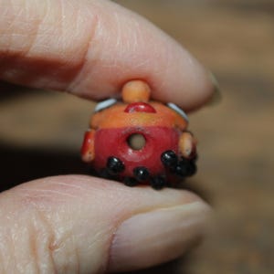 Może przedstawiać: Mała, ręcznie robiona szklana koralik w kolorze czerwonym i pomarańczowym. Koralik ma centralny otw&oacute;r i jest ozdobiony czarnymi kropkami i białymi akcentami. Ma małą, zaokrągloną g&oacute;rę i jest trzymany między palcami.