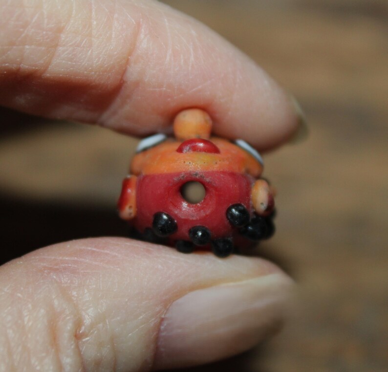 Może przedstawiać: Mała, ręcznie robiona szklana koralik w kolorze czerwonym i pomarańczowym. Koralik ma centralny otw&oacute;r i jest ozdobiony czarnymi kropkami i białymi akcentami. Ma małą, zaokrągloną g&oacute;rę i jest trzymany między palcami.