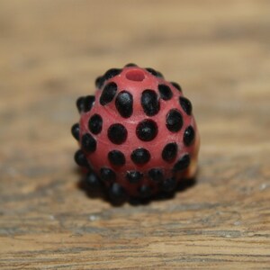 Może przedstawiać: Czerwony koralik z czarnymi kropkami i otworem u g&oacute;ry. Koralik ma teksturowaną powierzchnię i zaokrąglony kształt. Prawdopodobnie jest używany do tworzenia biżuterii lub innych rękodzieł.