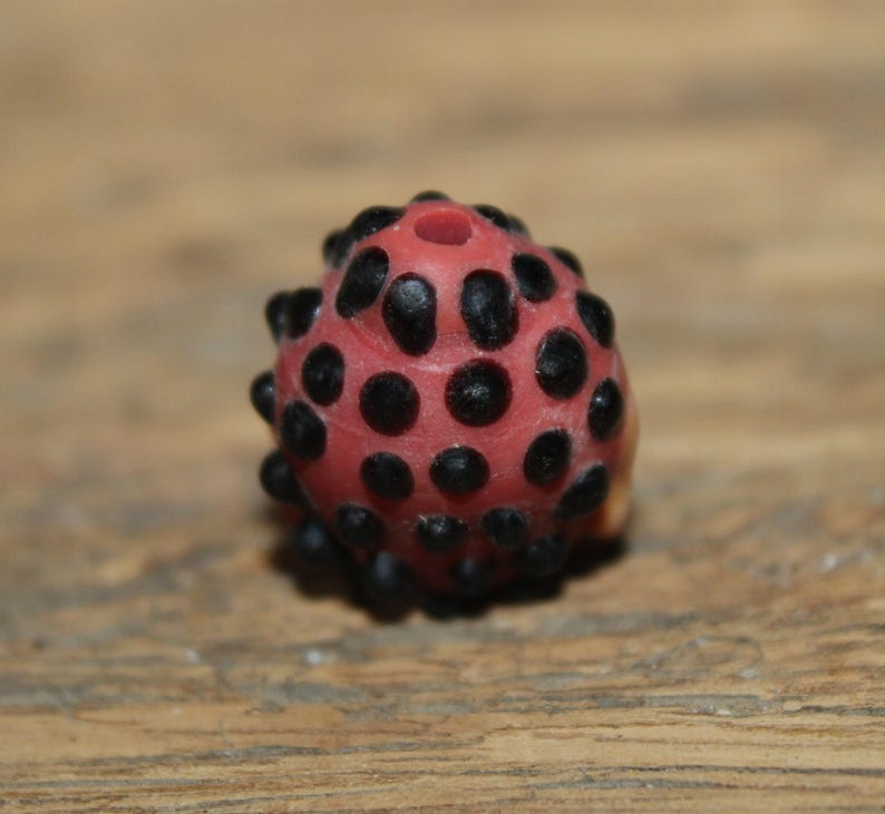 Może przedstawiać: Czerwony koralik z czarnymi kropkami i otworem u g&oacute;ry. Koralik ma teksturowaną powierzchnię i zaokrąglony kształt. Prawdopodobnie jest używany do tworzenia biżuterii lub innych rękodzieł.