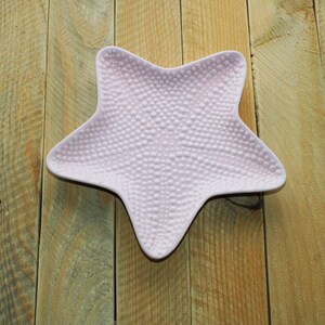 May include: A light pink, starfish-shaped ceramic dish with a textured surface. The dish has a dark brown rim and is set against a wooden background. This decorative dish is ideal for serving snacks or as a decorative accent.