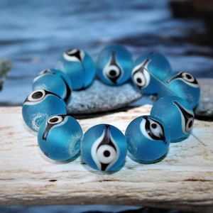 Könnte beinhalten: Eine Sammlung von mattierten, hellblauen Glasperlen. Jede Perle hat ein schwarz-weißes, augenähnliches Design. Die Perlen sind aufgefädelt, was darauf hindeutet, dass sie für die Schmuckherstellung oder das Basteln bestimmt sind. Der Hintergrund ist verschwommen blau und weiß.