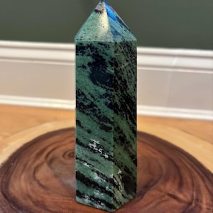 May include: A green and black stone obelisk with a pointed top. The polished stone has a rectangular shape and is set on a wooden surface. The obelisk is a decorative item.
