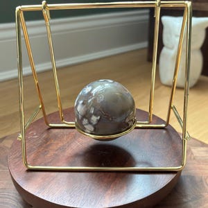 May include: A decorative gold metal stand cradles a spherical, patterned stone. The stone is a mix of gray, brown, and white. The stand is set on a round, dark wooden base, creating a floating effect.