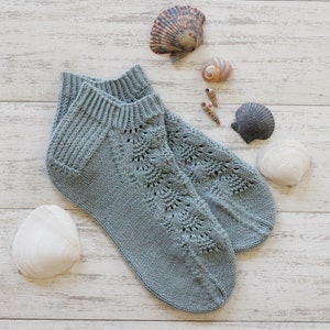 May include: A pair of grey knitted ankle socks with a lacy pattern. The socks are laid out on a white wooden surface with seashells scattered around them.