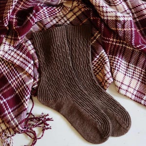 Pode incluir: Um par de meias de lã castanhas com um padrão de tricô de cabo. As meias estão sobre um tecido xadrez vermelho e branco.