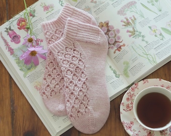 Könnte beinhalten: Ein Paar rosa Häkelsocken mit einem zarten Spitzenmuster. Die Socken liegen auf einem Buch mit botanischen Illustrationen.