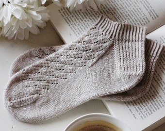 Könnte beinhalten: Hellgraue Strickstrümpfe mit Lochmuster liegen auf einem offenen Buch. Eine weiße Kaffeetasse und weiße Blumen ergänzen die Szene. Ein gemütliches Arrangement für Entspannung.