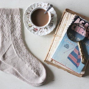 Peut inclure: Une paire de chaussettes en tricot beige avec un motif en dentelle, une tasse de café, un livre intitulé "Agatha Christie Miss Marple's Cases and Other Stories", et une loupe.