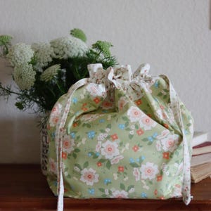 Könnte beinhalten: Ein salbeigrüner Beutel mit Kordelzug und einem wiederkehrenden Blumenmuster aus weißen, hellrosa und kleinen orangefarbenen Blüten. Der Beutel ist oben mit integrierten Stoffbändern gerafft, was einen voluminösen Rüscheneffekt am Rand erzeugt.
