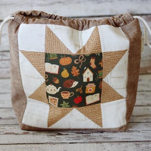 May include: A fabric drawstring pouch featuring a central star quilt block pattern. The star center displays a dark green background with small illustrations of pumpkins, pears, hedgehogs, houses, and tea cups. The pouch has brown side panels.